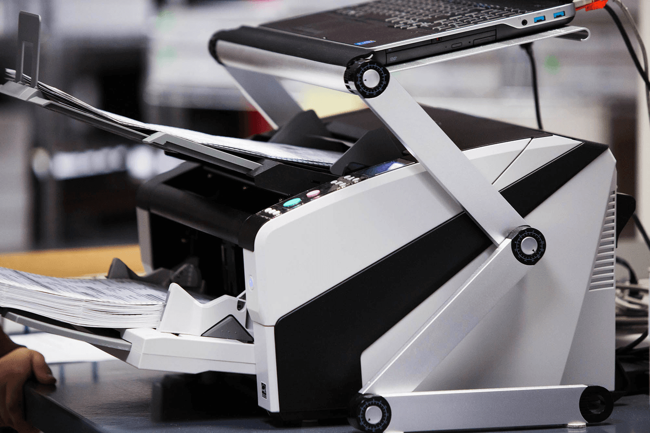 ClearCount central scanner with an open paper path, output ballots, and a laptop mounted above it